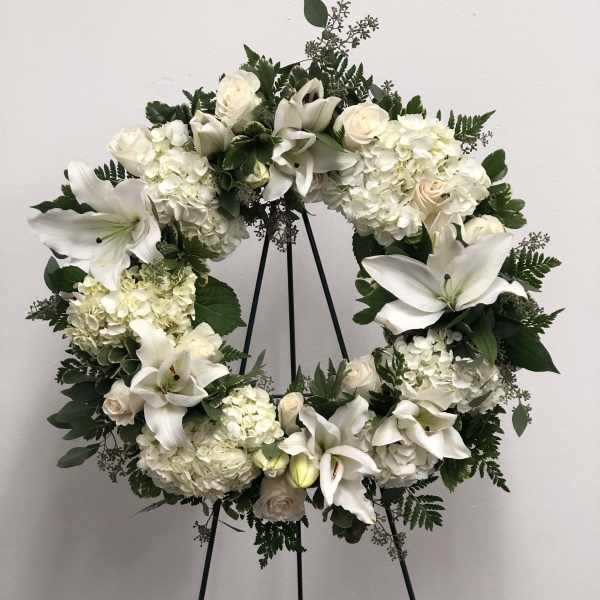 White floral wreath on a black stand with lilies, roses, and hydrangeas