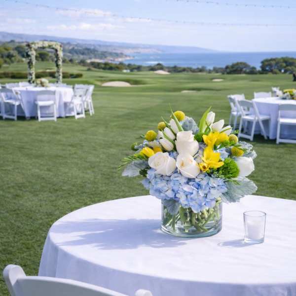 Low blue, white, and yellow flower centerpiece in a clear vase on an outdoor table with white linens