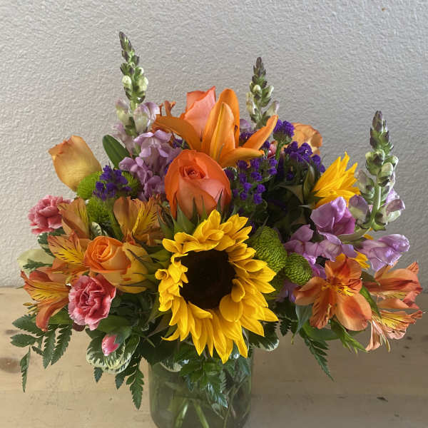 Colorful bouquet with sunflowers, roses, lilies, and purple filler in a glass vase