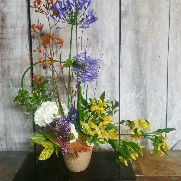 Tall mixed floral arrangement in a beige vase with purple, yellow, white, and orange blooms.