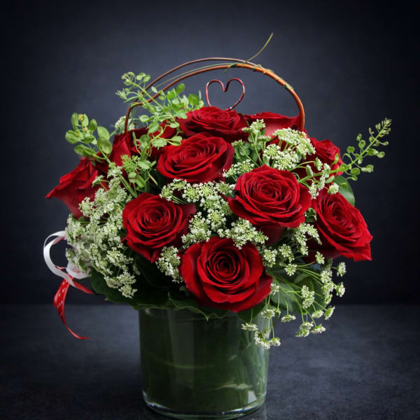 Red roses arranged in a glass vase with white filler flowers and a heart pick
