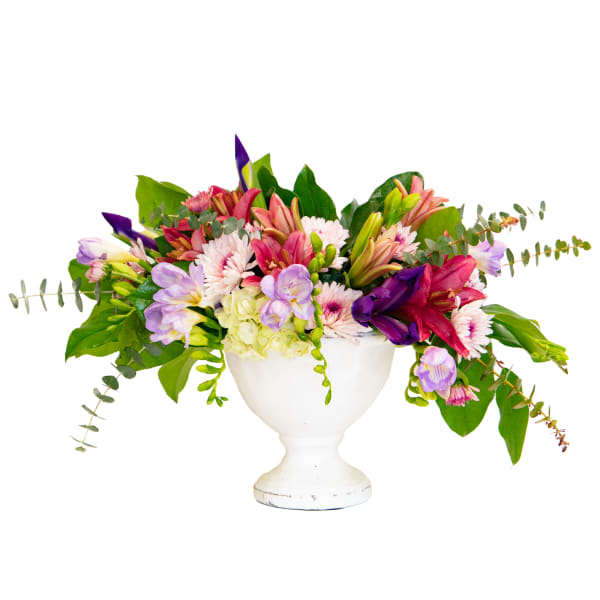 Low arrangement of pink lilies, daisies, and purple flowers in a white pedestal bowl