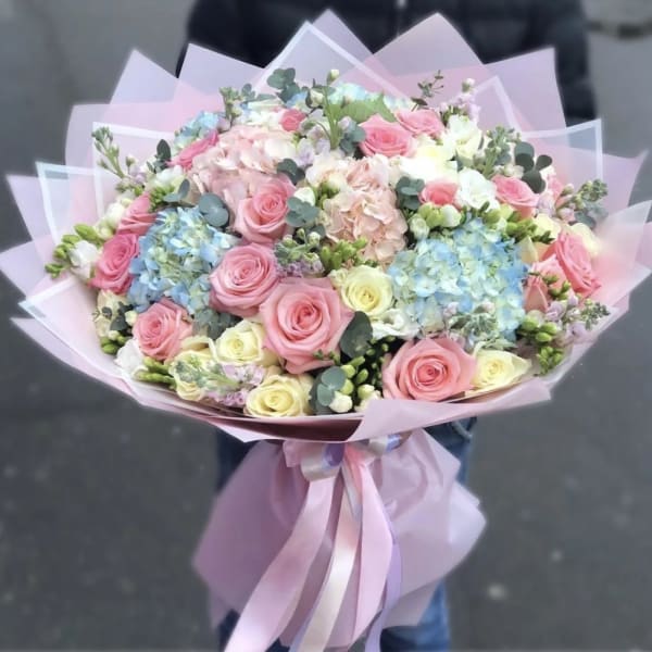 Large pastel bouquet of pink and cream roses with blue hydrangeas wrapped in pink paper and ribbon