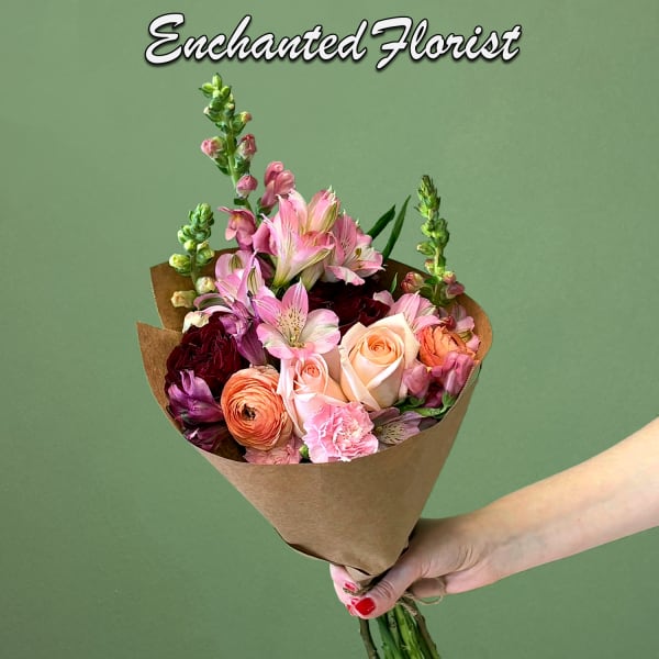 Hand-tied bouquet of pink and peach roses, ranunculus, and mixed blooms wrapped in brown paper.