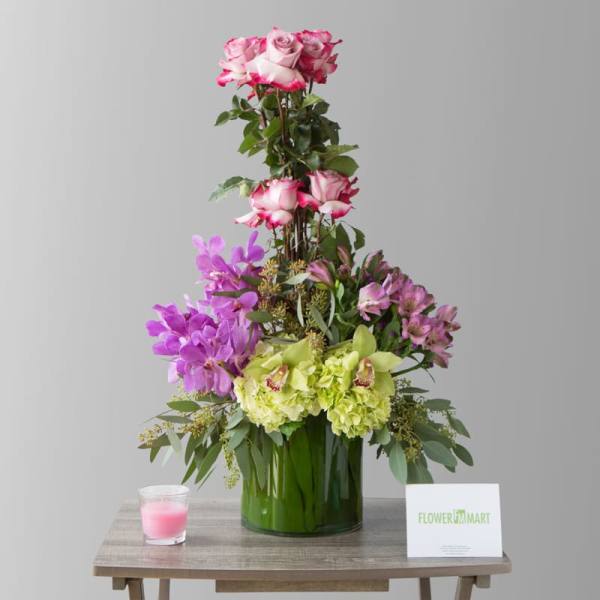 Tall floral arrangement with pink roses, purple orchids, and green hydrangeas in a glass vase