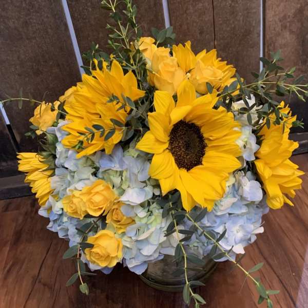 Yellow sunflowers and roses in a low glass vase with pale blue hydrangeas.