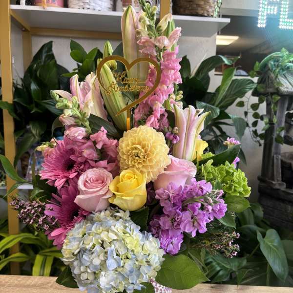 Mixed pink and yellow flower arrangement in a pink vase with a "Happy Anniversary" topper