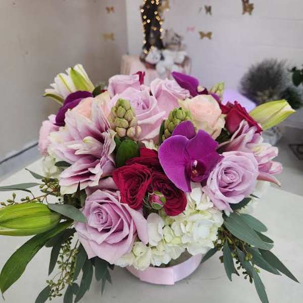 Low round arrangement of pink and purple roses, orchids, and white hydrangea in a hatbox container