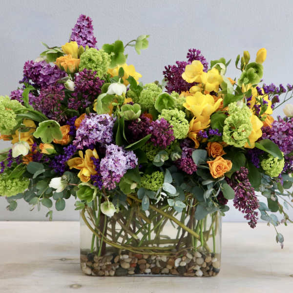 Lush mixed arrangement of purple, yellow, green, and orange flowers in a rectangular glass vase with pebbles.