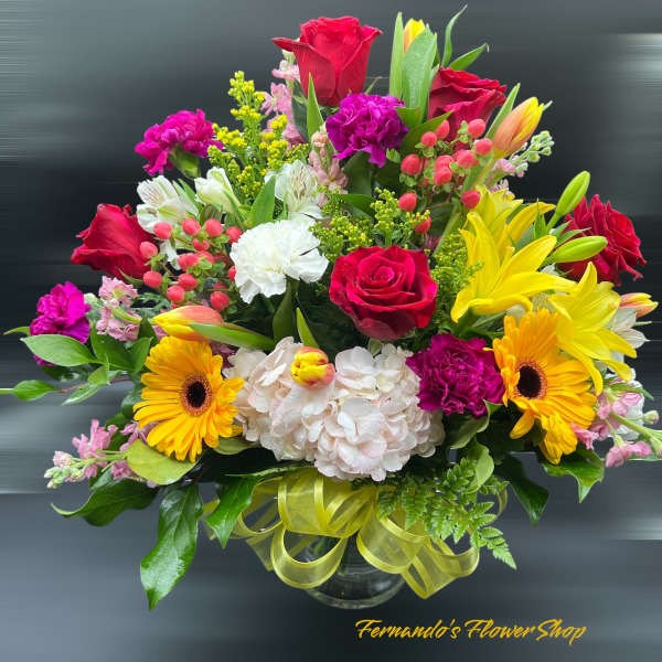 Bright mixed flower arrangement with red roses, yellow lilies, and gerbera daisies in a glass vase.