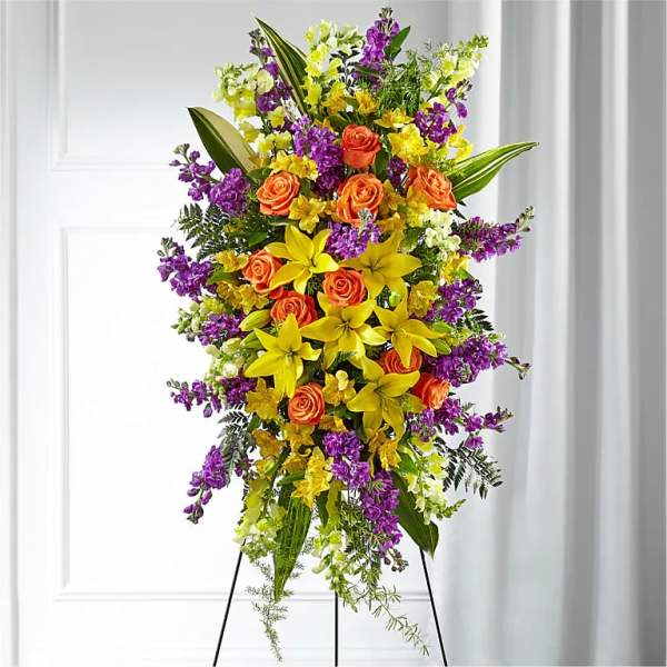 Tall standing spray with yellow lilies, orange roses, and purple and white flowers on a metal easel.