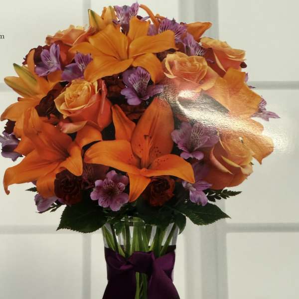Orange lilies and roses in a clear vase with a purple ribbon