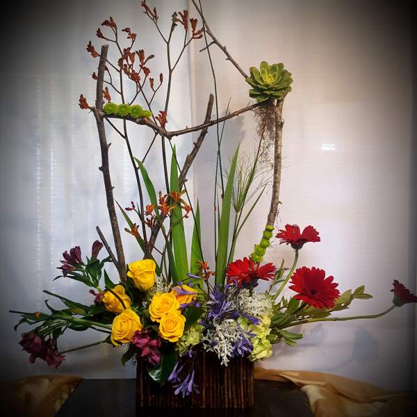 Tall floral arrangement with yellow roses, red daisies, and branches in a dark vase