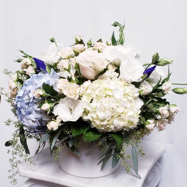 Low arrangement of white and blue hydrangeas, roses, and lisianthus in a round white container