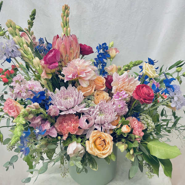 Large mixed floral arrangement in a pale green vase with pink, peach, blue, and lavender blooms.