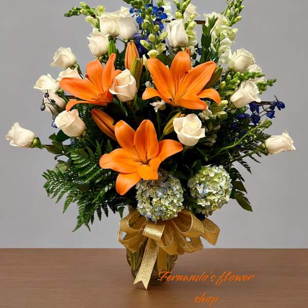 Tall arrangement of orange lilies, white roses, blue flowers, and hydrangeas in a gold vase with a bow
