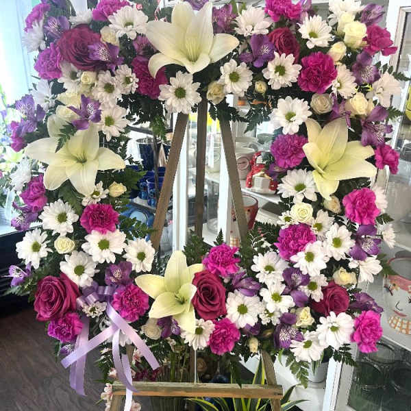 Large floral wreath on an easel with white lilies, daisies, roses, and purple blooms
