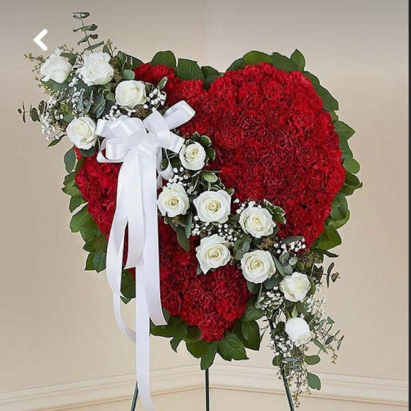 Heart-shaped red carnation standing spray with white roses, white ribbon bow, and greenery on an easel.