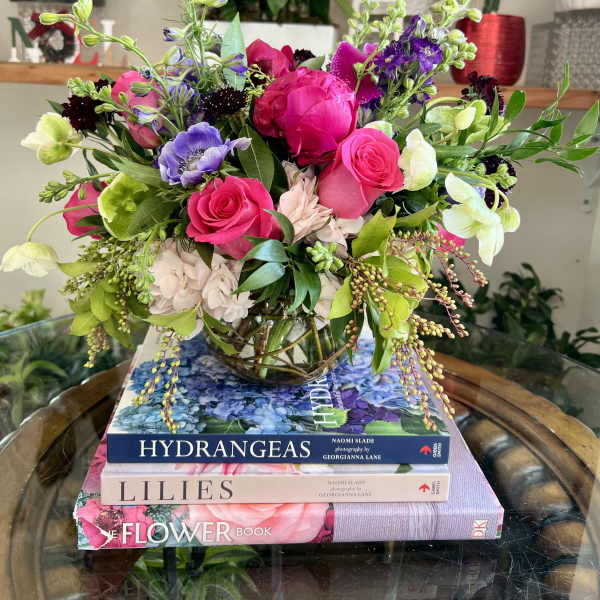 Low glass vase of bright pink roses and mixed purple and green flowers on a stack of flower books.