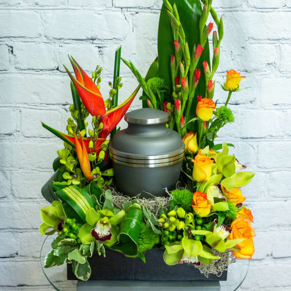 Cremation urn surrounded by orange roses, green orchids, and tropical flowers in a tall arrangement