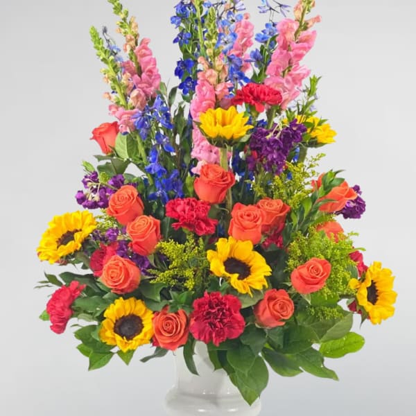 Colorful mixed bouquet in a clear glass vase with roses, sunflowers, and tall blue blooms