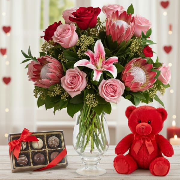 Pink and red bouquet with lilies and protea in a glass vase beside chocolates and a red teddy bear