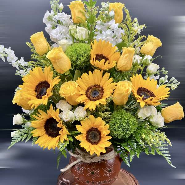 Arrangement of yellow sunflowers and roses with white blooms in a rustic clay pot on a wood slab base