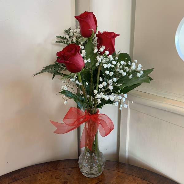 Three red roses in a clear glass vase with baby's breath and a red ribbon
