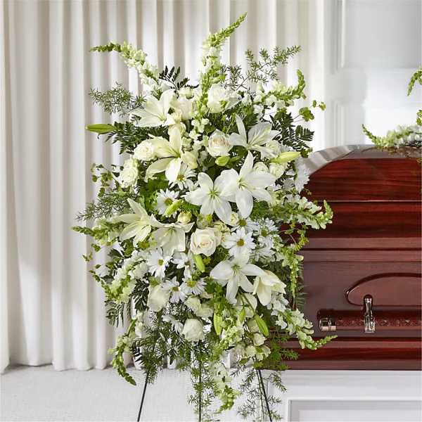 Standing spray of white lilies, roses, and daisies on an easel beside a wooden casket.