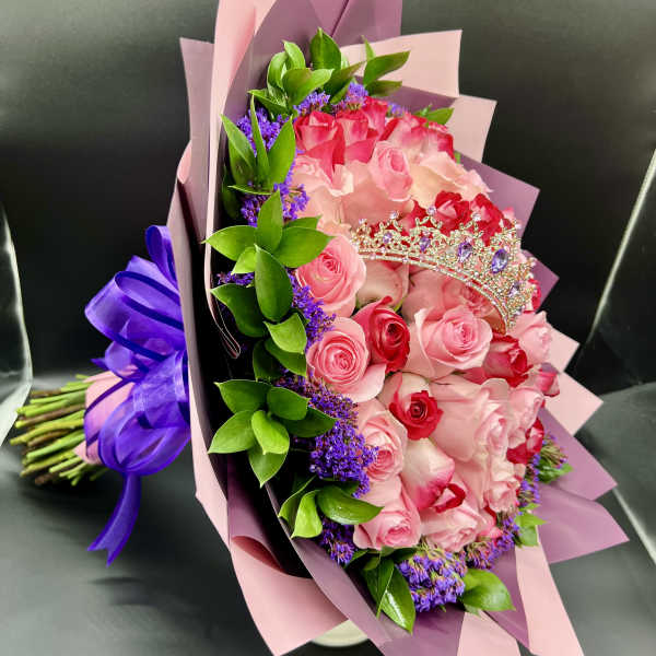 Bouquet of pink and red roses with purple filler flowers, wrapped in pink paper with a small tiara decoration.