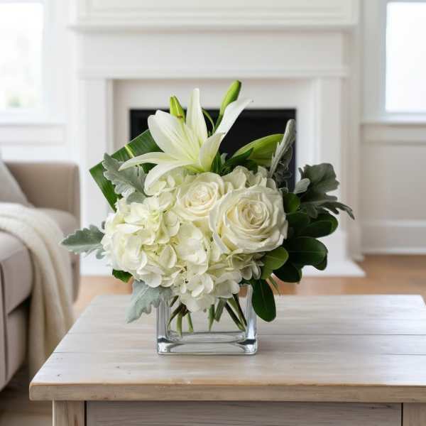Low white arrangement of lilies, roses, and hydrangeas in a clear glass cube vase on a light wood table