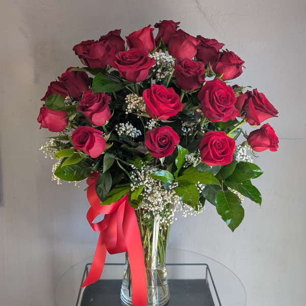 Tall arrangement of red roses with white filler flowers in a clear vase with a red ribbon bow