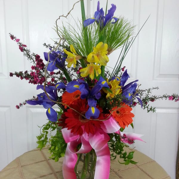 Colorful bouquet with iris, gerbera daisies, and a pink ribbon in a glass vase