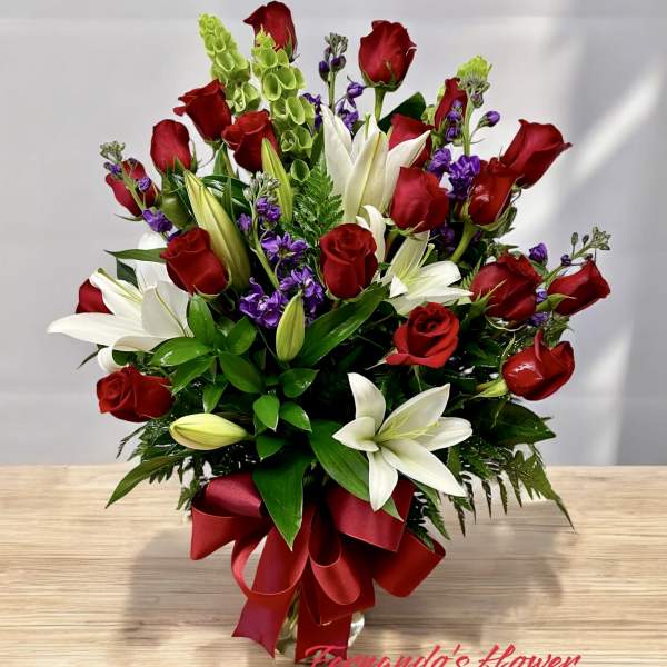 Tall arrangement of red roses, white lilies, purple flowers and green bells in a glass vase with red bow