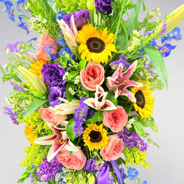 Tall standing floral spray with sunflowers, pink roses, lilies, and purple flowers on an easel