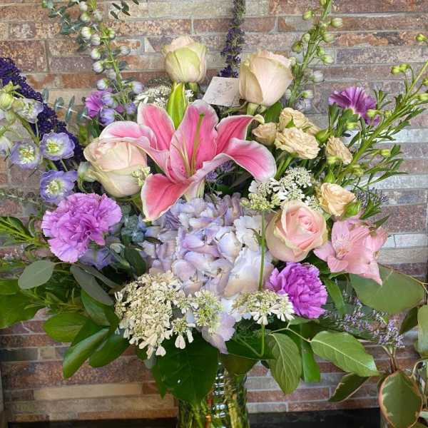 Mixed pastel bouquet with lilies, roses, hydrangea, and purple accents in a glass vase