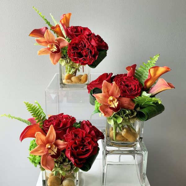Three compact red and orange floral arrangements in glass cube vases with river rocks