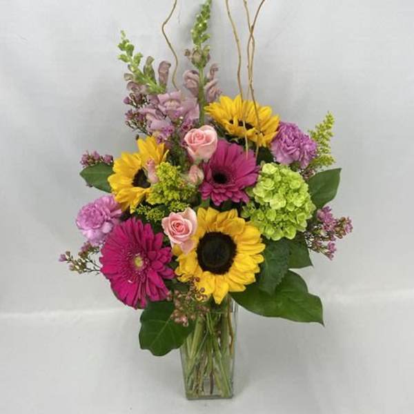 Tall mixed arrangement with sunflowers, pink gerberas, roses, and green hydrangeas in a clear glass vase