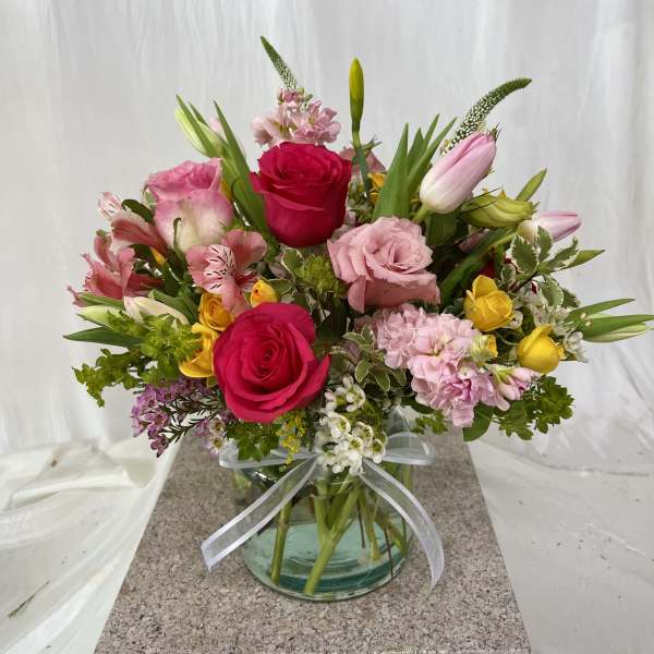 Mixed pink, yellow, and white flowers arranged in a glass vase with a ribbon