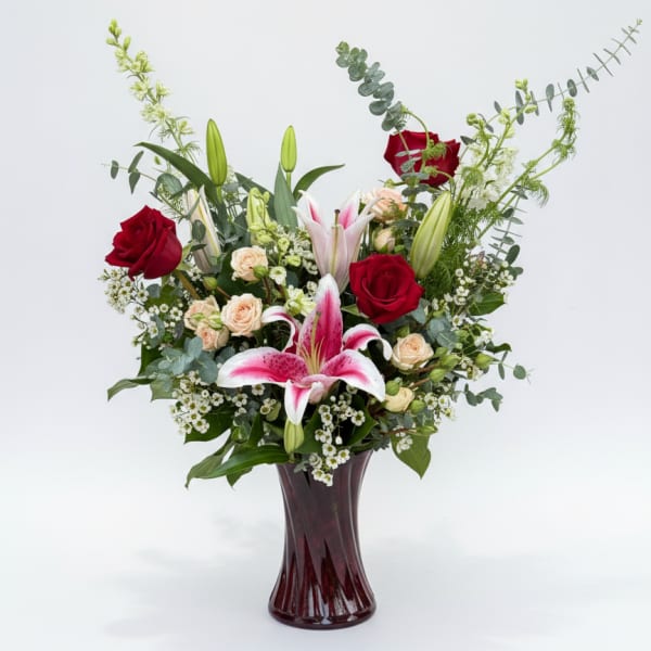 Tall arrangement of red roses, pink lilies, and peach spray roses in a dark red glass vase
