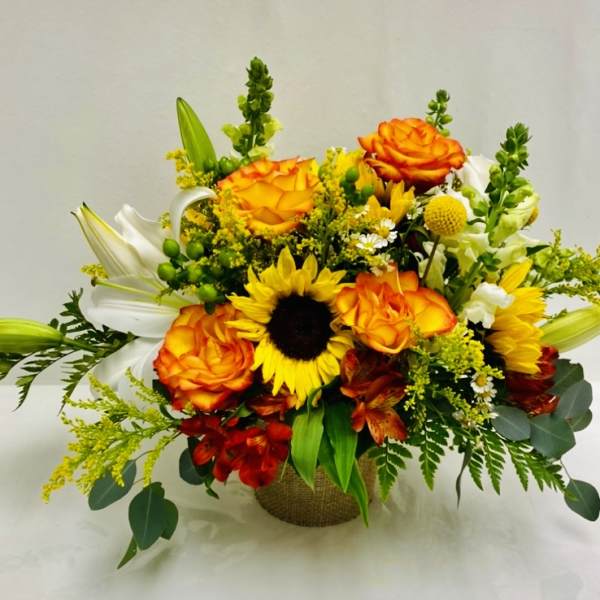 Bouquet of orange roses, sunflowers, and white lilies in a burlap-wrapped container