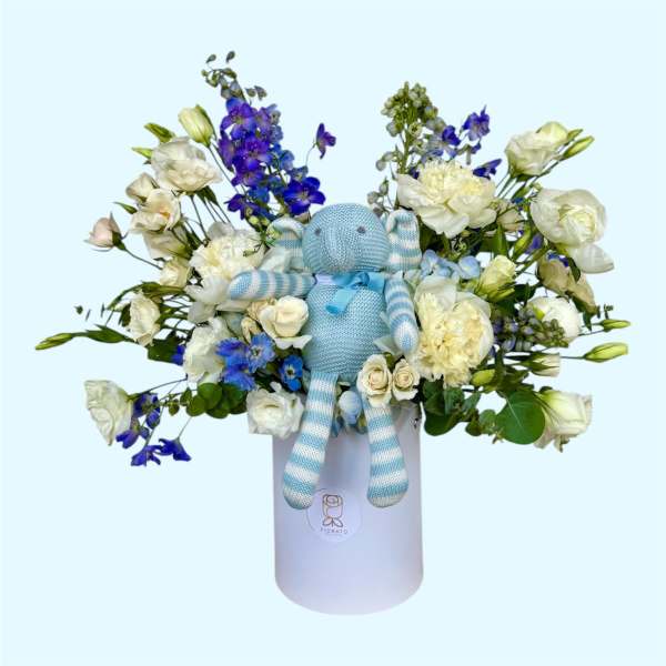 Blue teddy bear in a white box surrounded by white and blue flowers