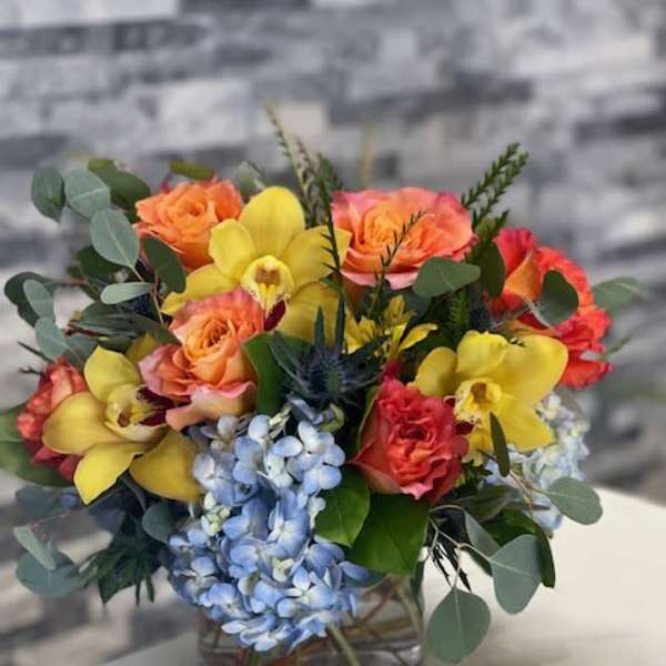 Short arrangement of orange roses, yellow orchids, and blue hydrangeas in a clear glass vase.