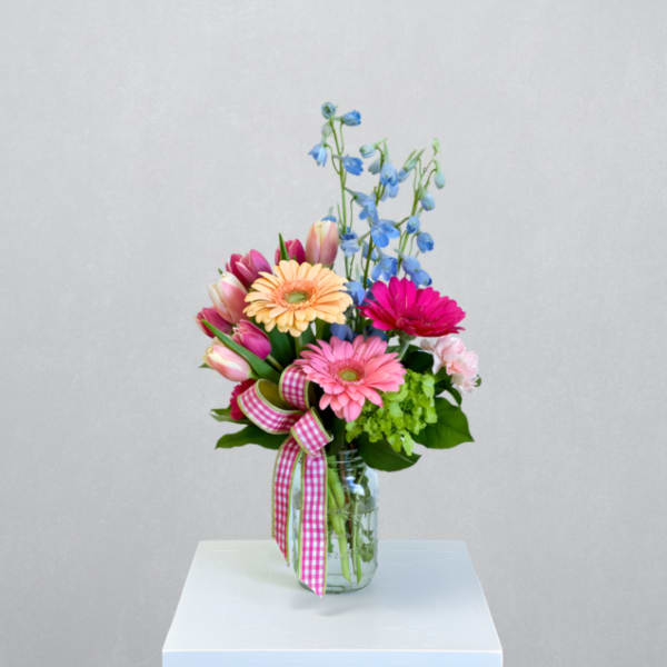 Mixed pink gerbera daisies, tulips, and blue delphinium in a mason jar with gingham ribbon