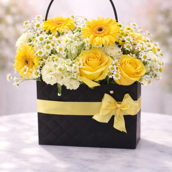 Yellow roses and gerbera daisies with white daisies in a black gift bag with yellow ribbon.
