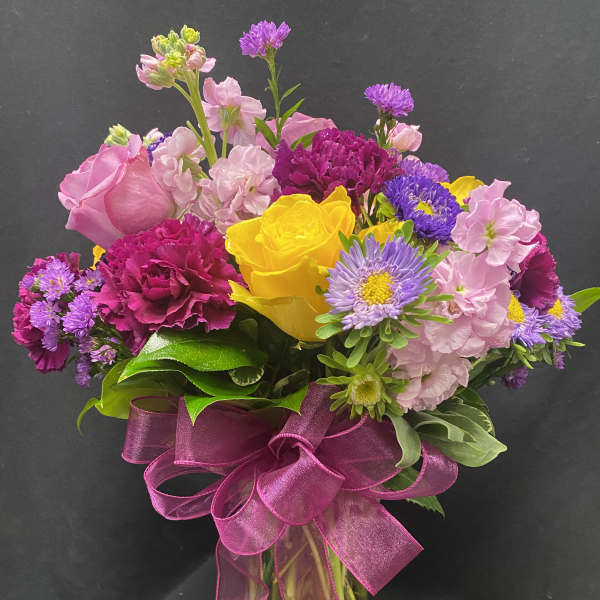 Mixed bouquet of yellow roses, pink stock, and purple asters in a clear vase with a magenta bow.
