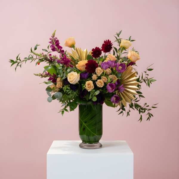 Tall peach, burgundy, and purple flower arrangement with gold accents in a green-filled glass vase.