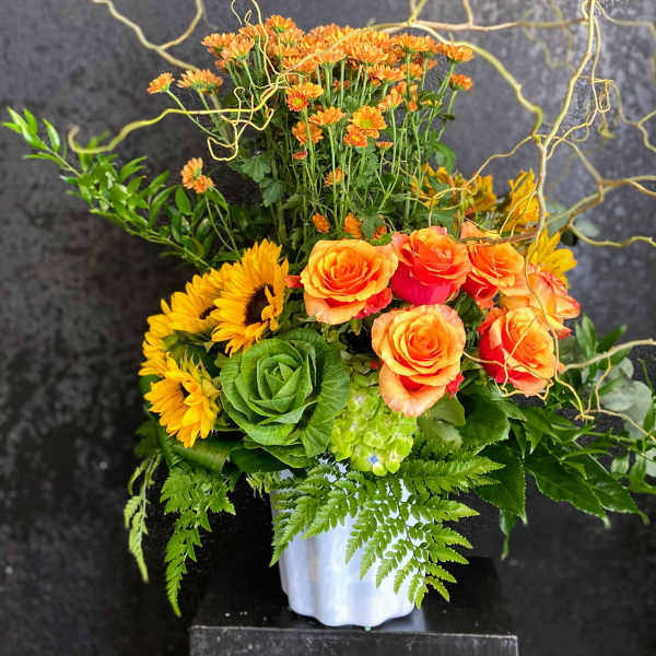 Tall orange and yellow arrangement with roses, sunflowers and mums in a white vase with curly branches