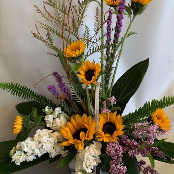 Tall floral arrangement with sunflowers, white blooms, and purple flowers in a square container