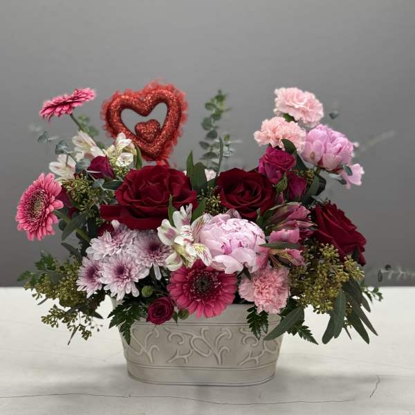Pink and red mixed flower arrangement in a decorative white container with a glitter heart pick